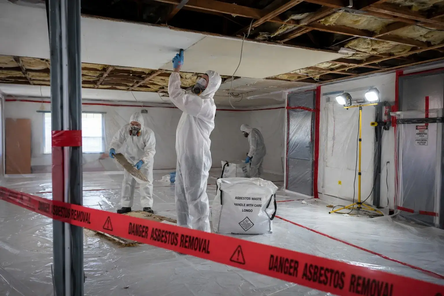 Indoor asbestos removal in a containment zone with full PPE and hazard warning tape