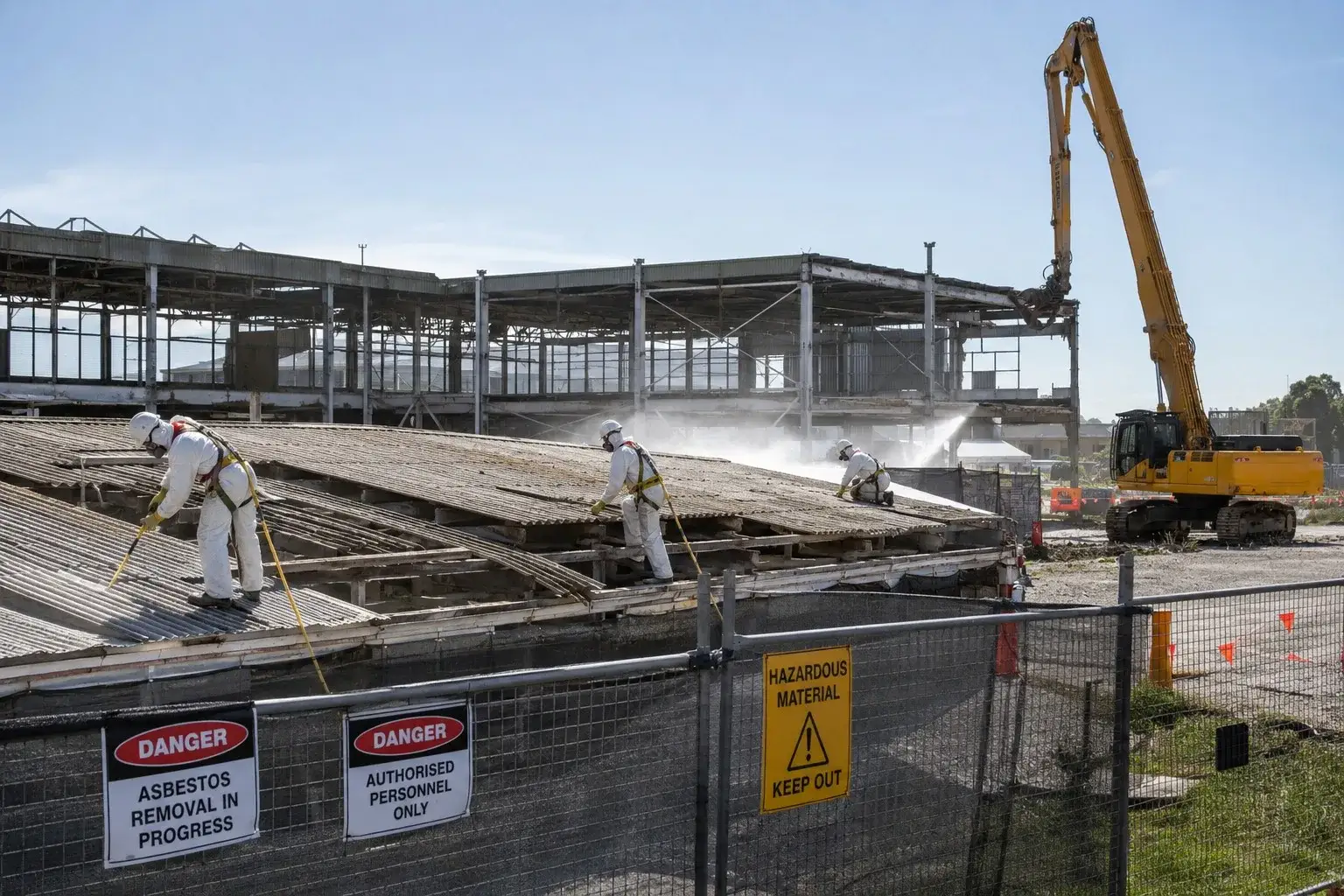 Workers in PPE removing asbestos from a large industrial roof in Adelaide with excavator and danger signs